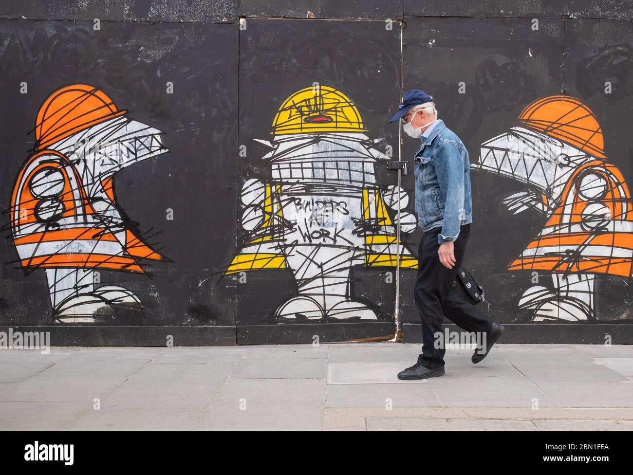 A man wearing a protective face mask walks past a street art mural showing construction workers in central London, as Prime Minister Boris Johnson said people who cannot work from home should be 'actively encouraged' to return to their jobs. Stock Photo