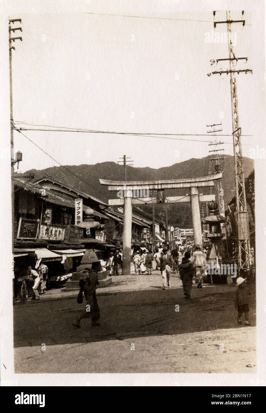 [ 1930s Japan - Sacred Torii Gate in Shopping Street in Kobe ] —   Stone lanterns and a scared torii gate at the entrance way to Ikuta Jinja, a Shinto shrine in Kobe, Hyogo Prefecture, ca 1932 (Showa 7).  Lots of shops line the street. The shrine is among the oldest shrines in Japan and is mentioned in the Nihon Shoki, the second oldest book of classical Japanese history.  The Battle of Ichi-no-Tani (1184) took place in and around Ikuta Shrine.  20th century gelatin silver print. Stock Photo