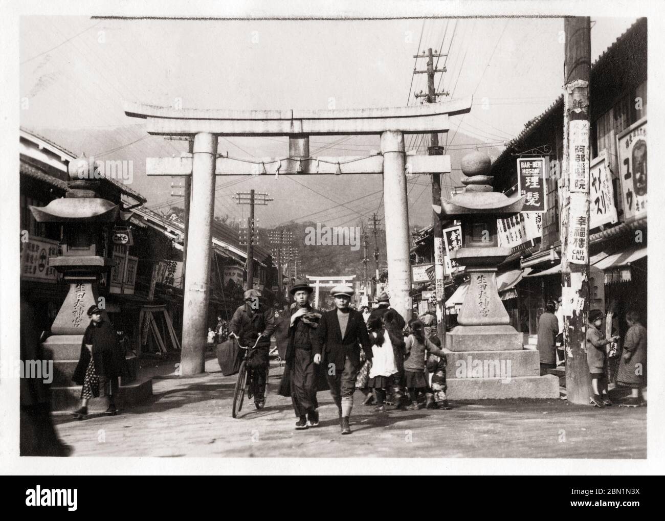 [ 1930s Japan - Sacred Torii Gate in Shopping Street in Kobe ] —   Stone lanterns and a scared torii gate at the entrance way to Ikuta Jinja, a Shinto shrine in Kobe, Hyogo Prefecture, ca 1932 (Showa 7).  Lots of shops line the street. The shrine is among the oldest shrines in Japan and is mentioned in the Nihon Shoki, the second oldest book of classical Japanese history.  The Battle of Ichi-no-Tani (1184) took place in and around Ikuta Shrine.  20th century gelatin silver print. Stock Photo
