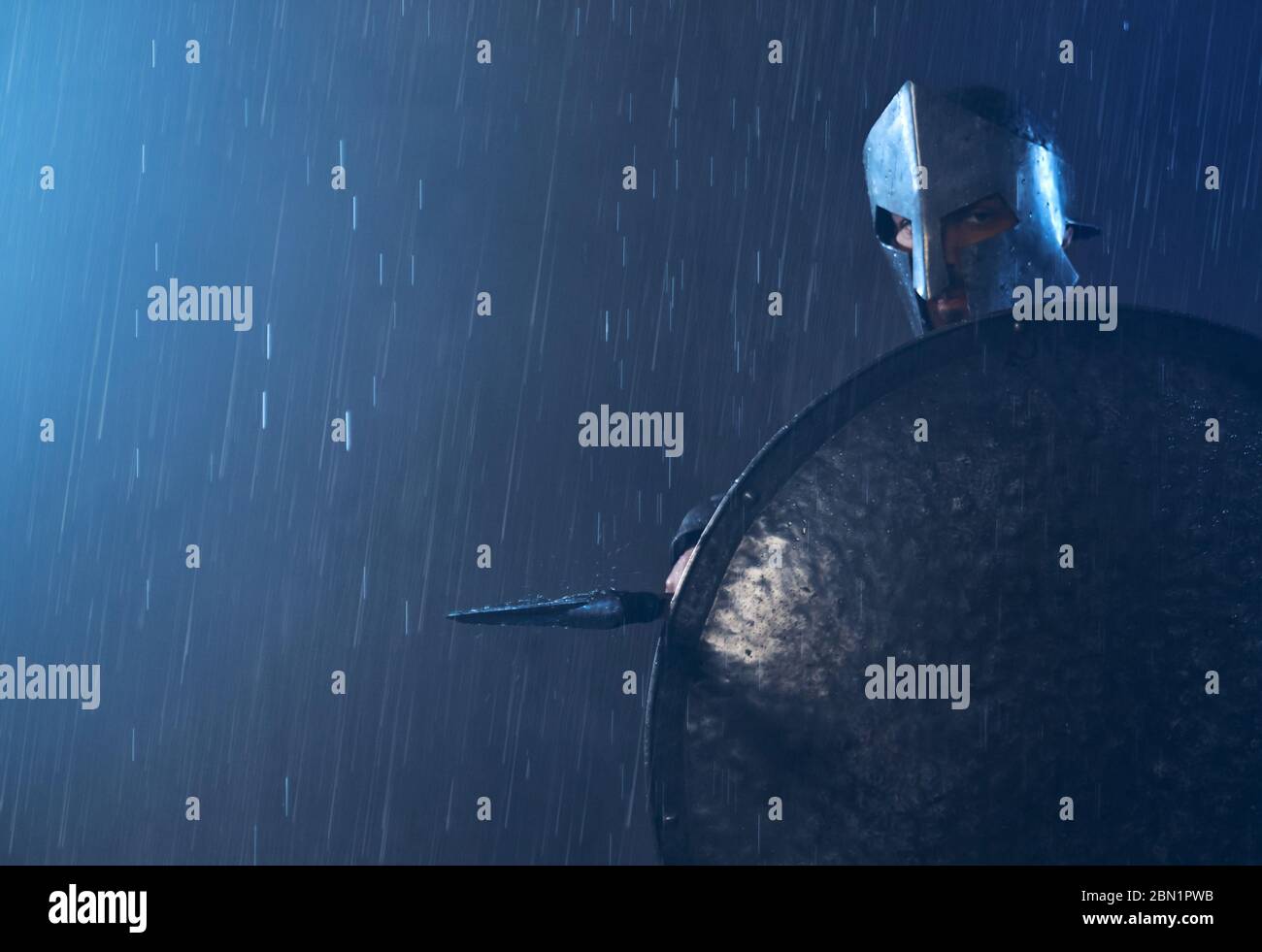 Portrait of spartan standing outdoors with spear and looking at camera. Front view of man in helmet hiding behind iron shield and pointing weapon in bad cloudy rainy weather. Ancient sparta concept. Stock Photo
