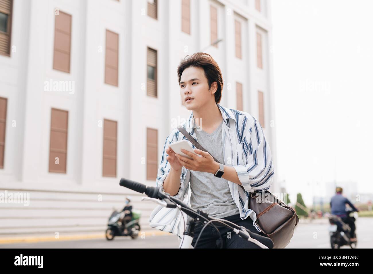 Stylish man chatting while using mobile phone while sitting on bicycle, outdoors. Dressed up in plaid shirt, t-shirt and jeans. Close-up. Stock Photo