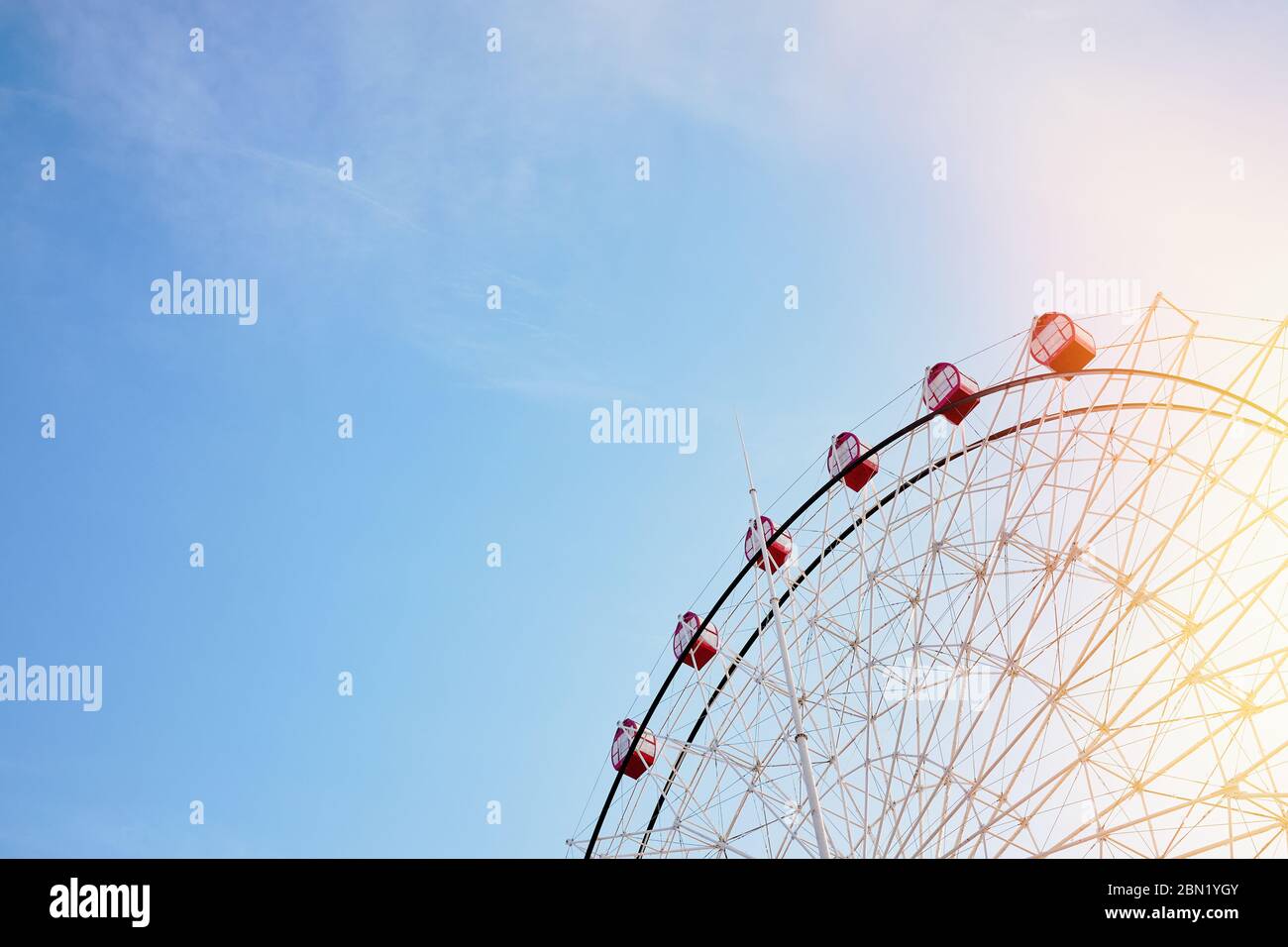 Ferris wheel with bright red booths on background of serene clear blue sky with sunflare as symbol of joy recreation relaxation fun and good time, wit Stock Photo