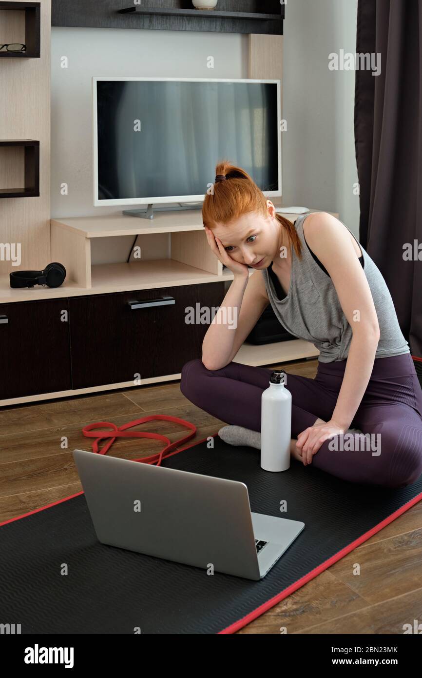 The girl sits on a sports mat and backs her head with her hand shocked looking into the laptop.  Stock Photo