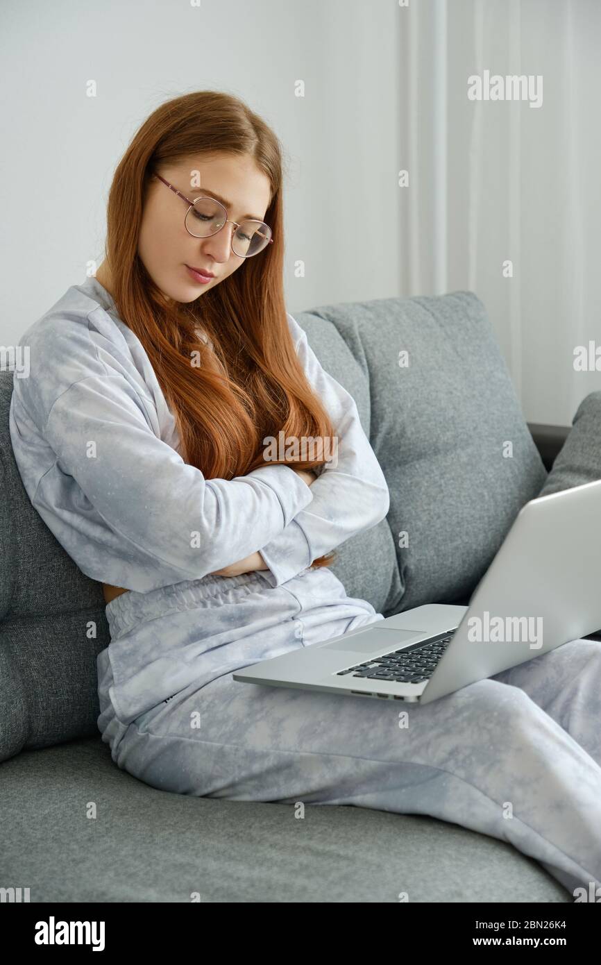 A red-haired girl in glasses and pajamas sits on the couch with a laptop, arms folded and her eyes closed.  Stock Photo