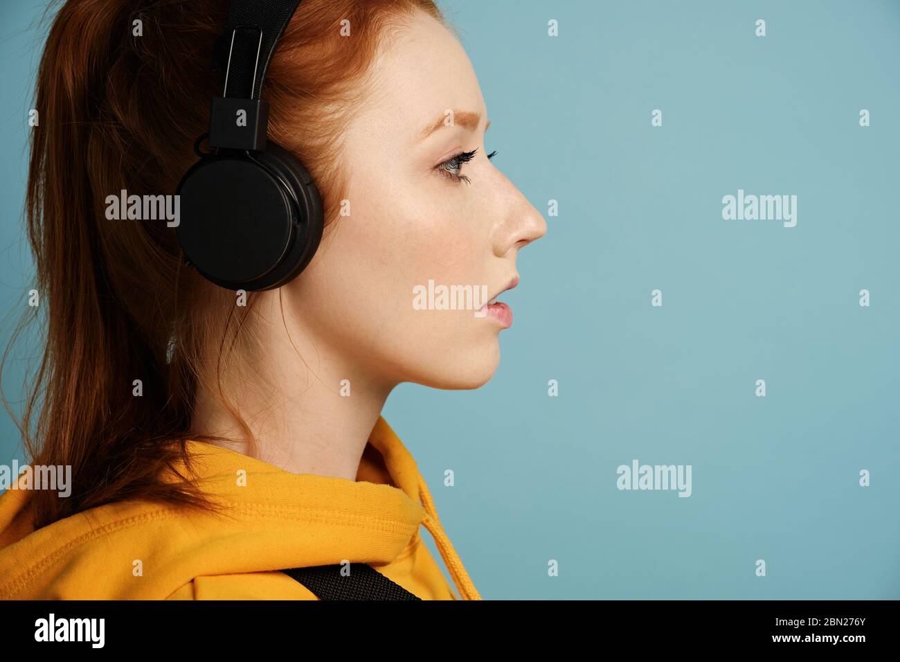 Head shot. A red-haired girl wearing headphones and a yellow hoodie stands in profile on a blue background Stock Photo