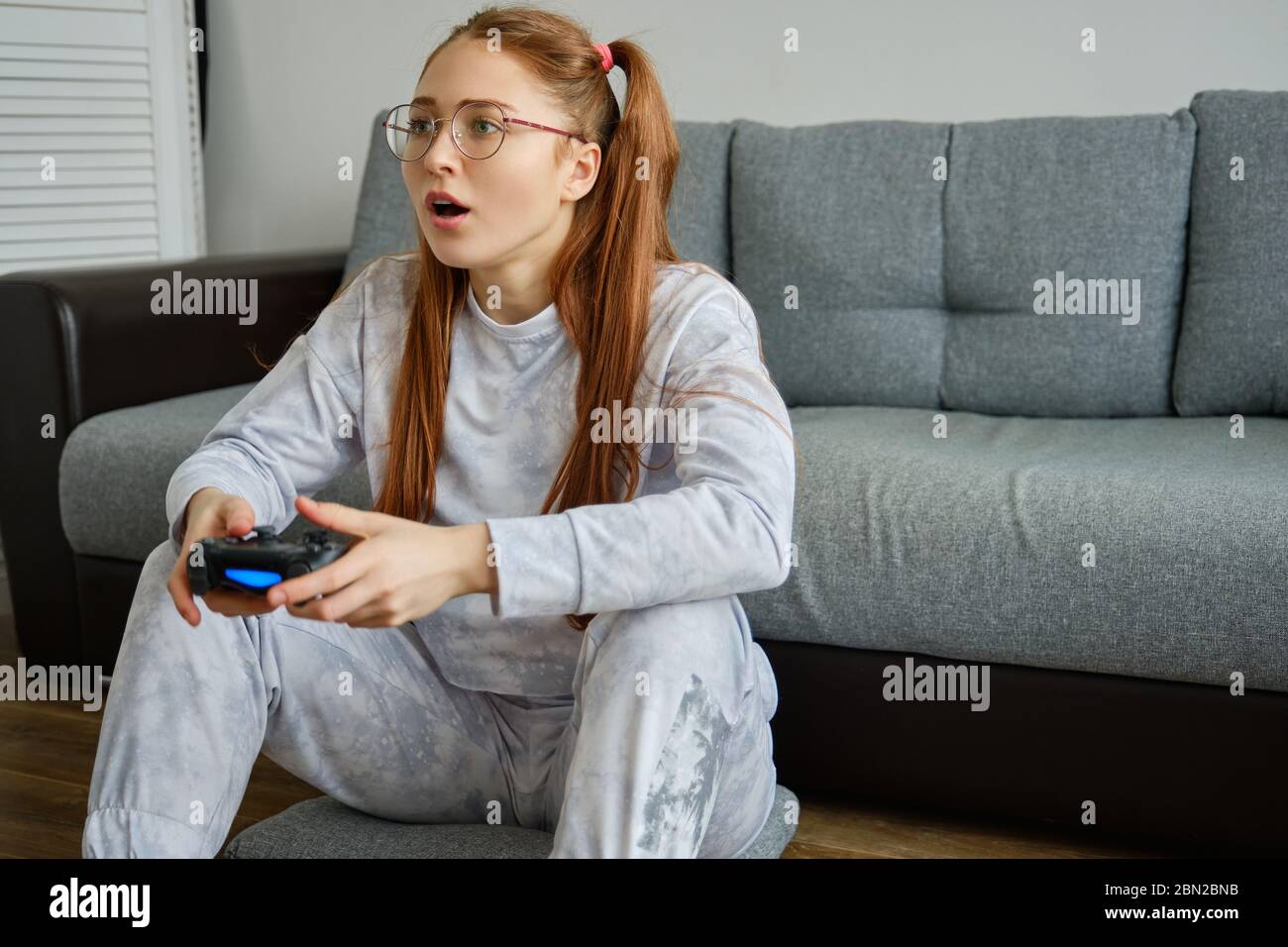 Red girl with tails in glasses sits on the floor and enthusiastically plays joystick. Stock Photo