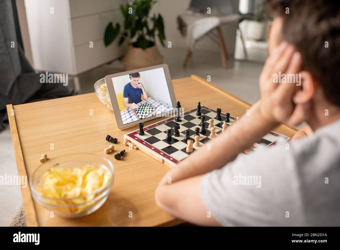 Over shoulder view of man sitting at coffee table and deciding about chess move while playing with friend via video chat Stock Photo