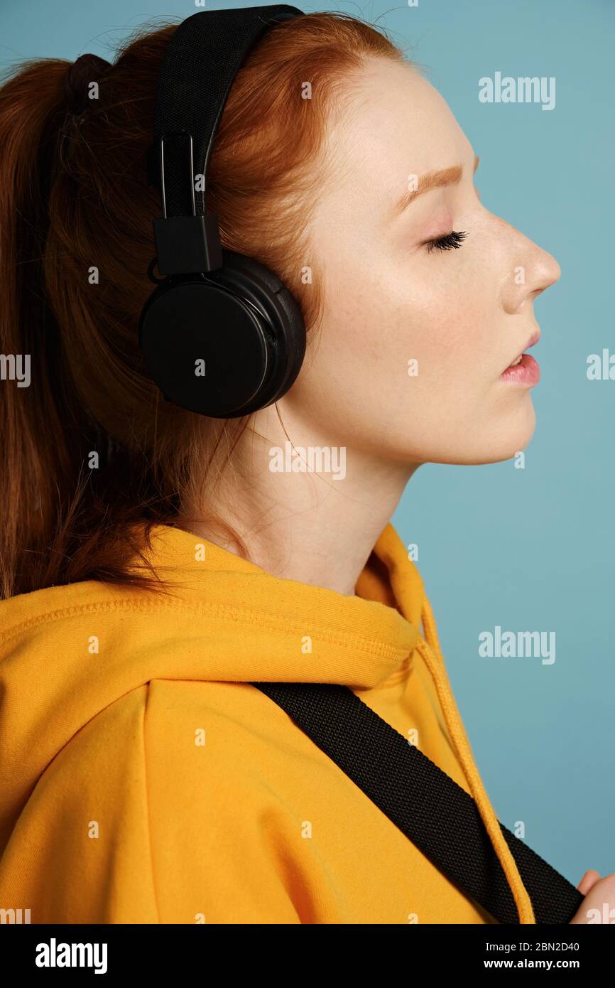 Head shot. A red-haired girl wearing headphones and a yellow hoodie stands on a blue background in a profile with her eyes closed. Stock Photo