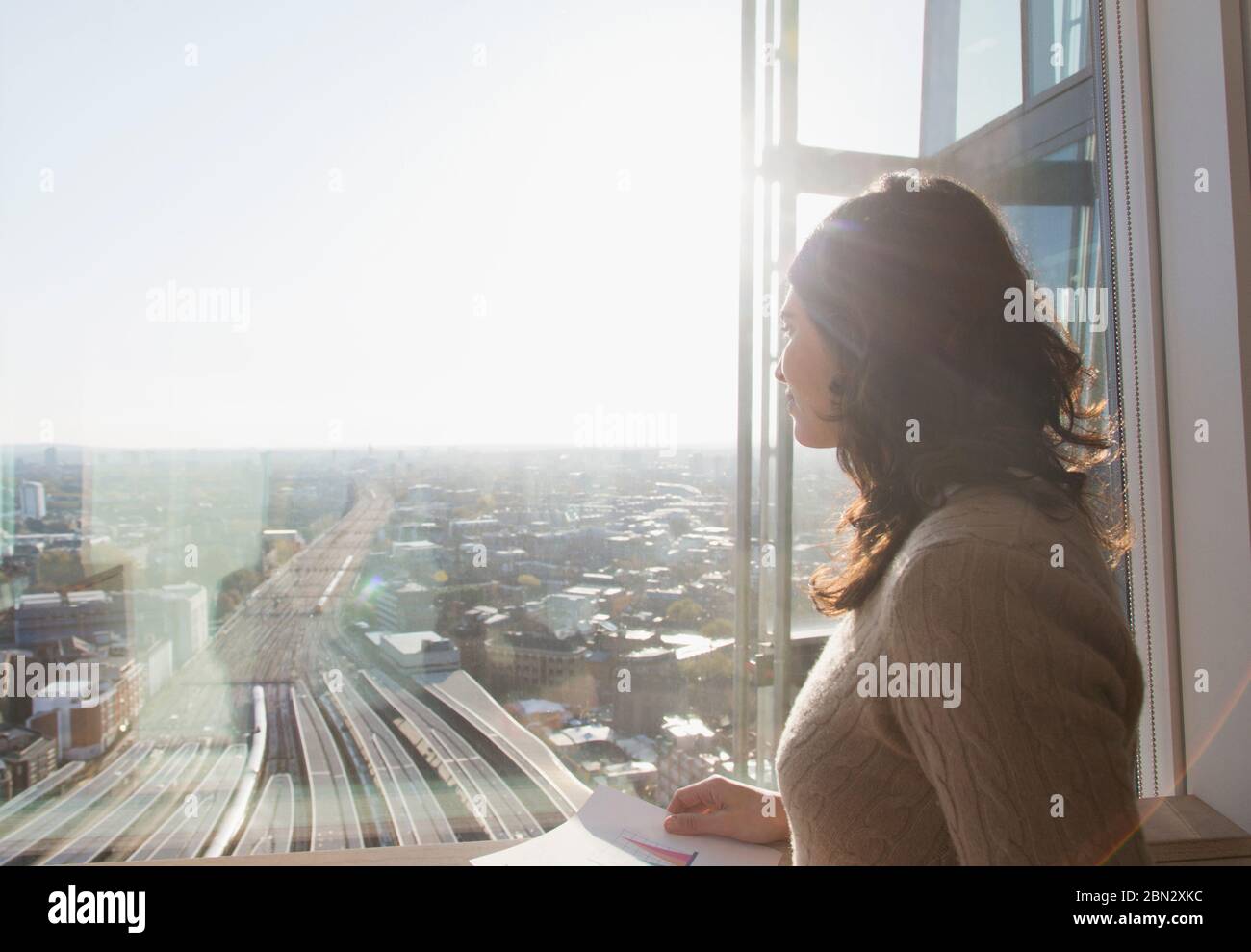 Thoughtful businesswoman at sunny window overlooking city Stock Photo
