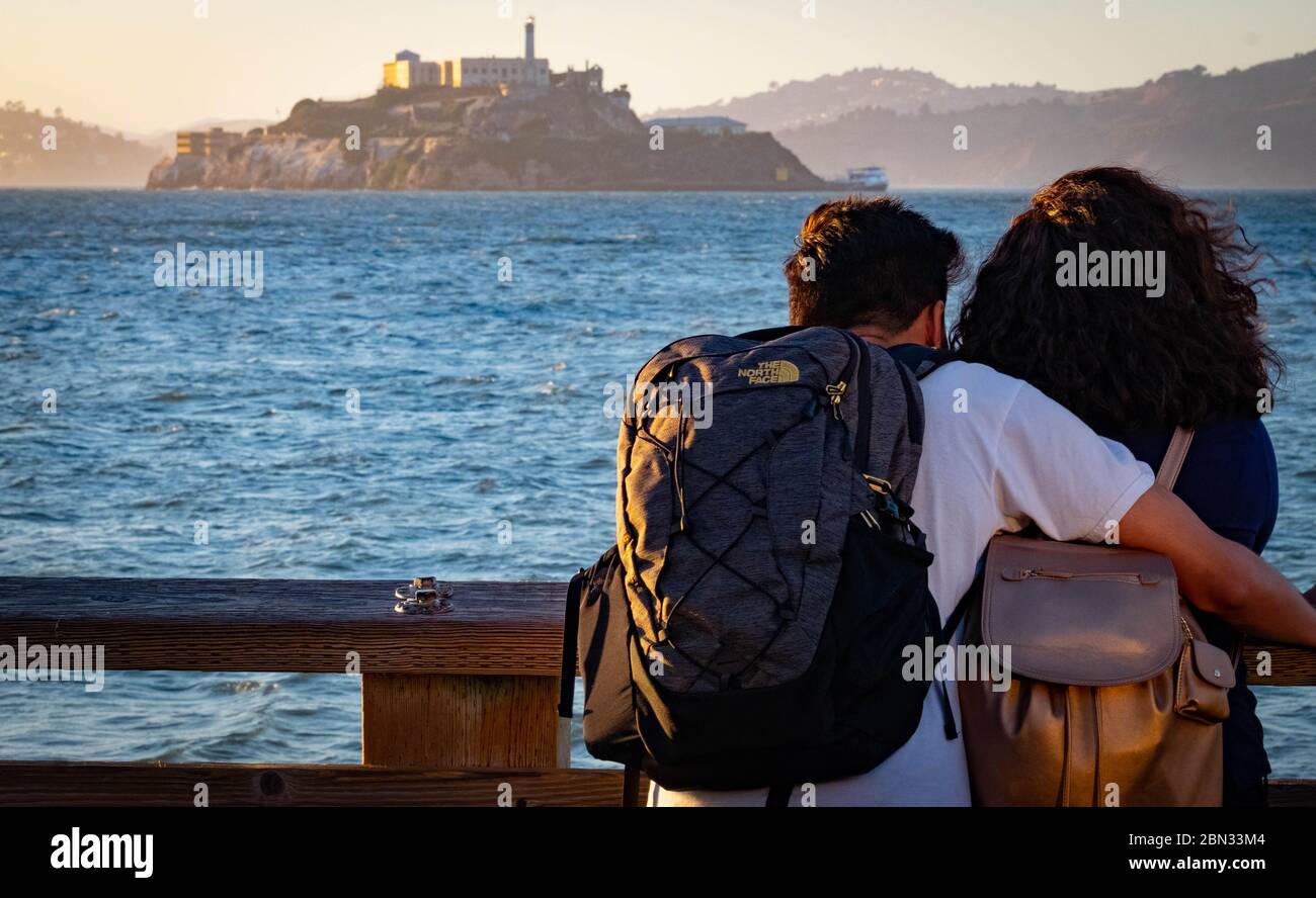 A young couple standing at the rail on Pier 39, Fishermen's Walk in San Francisco gazing out across the bay to Alcatraz Prison as the sun sets. Stock Photo