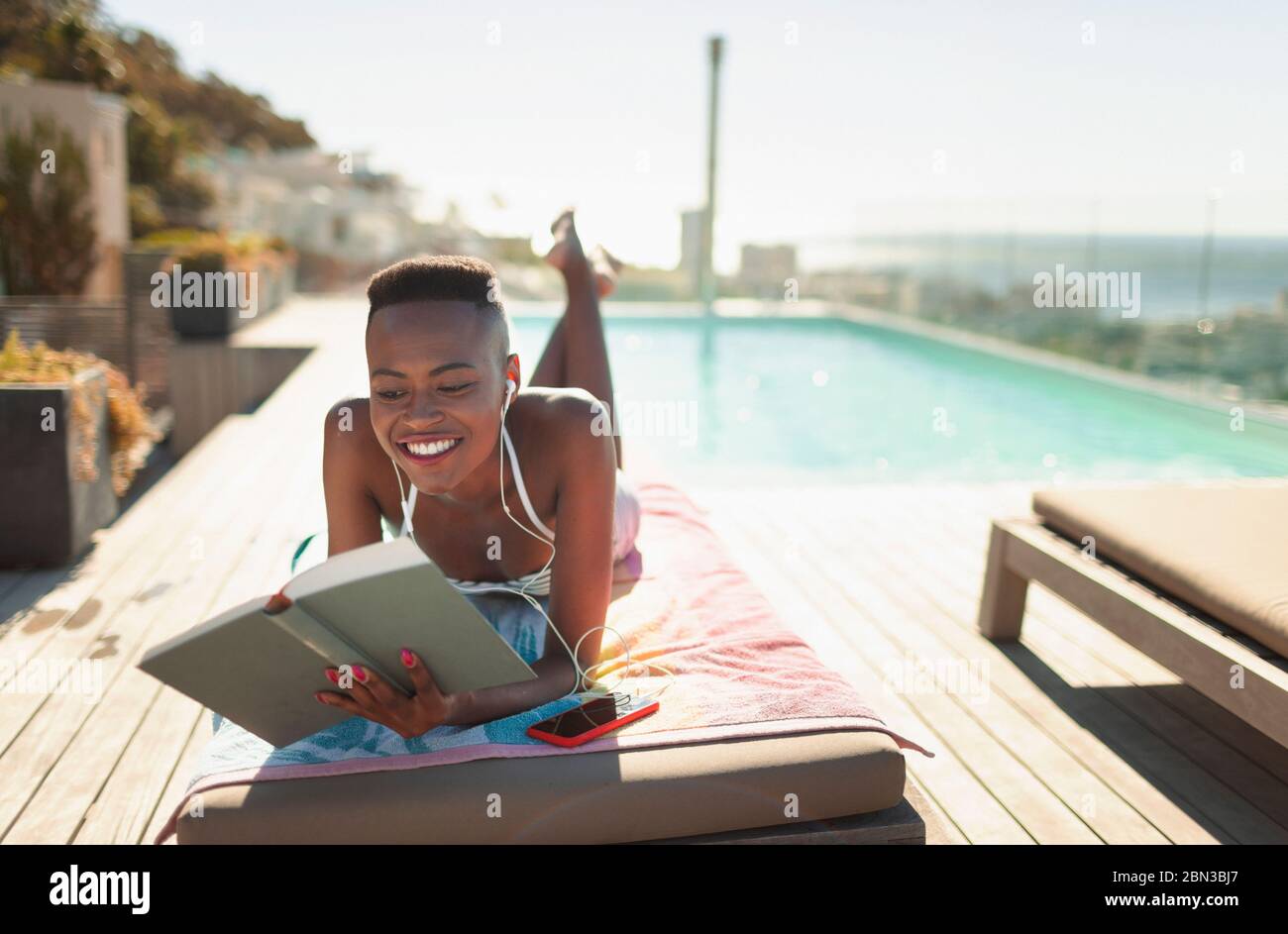 Smiling, carefree young woman reading book at sunny poolside Stock Photo