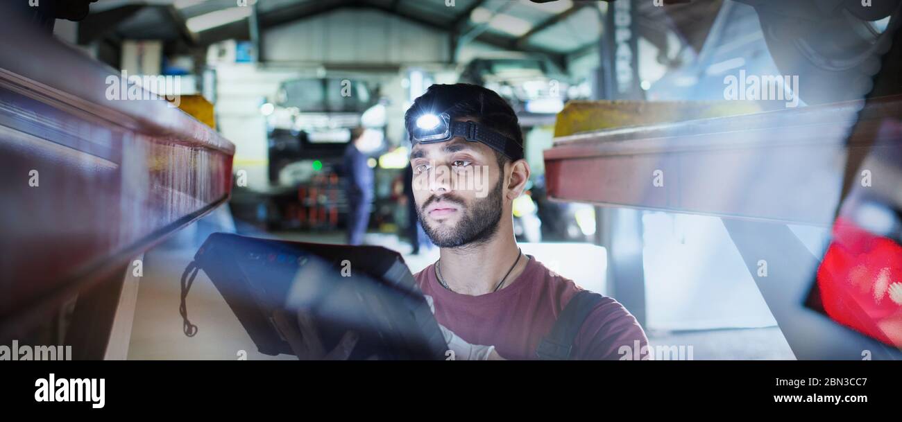 Focused male mechanic with headlight and diagnostic equipment working under car in auto repair shop Stock Photo