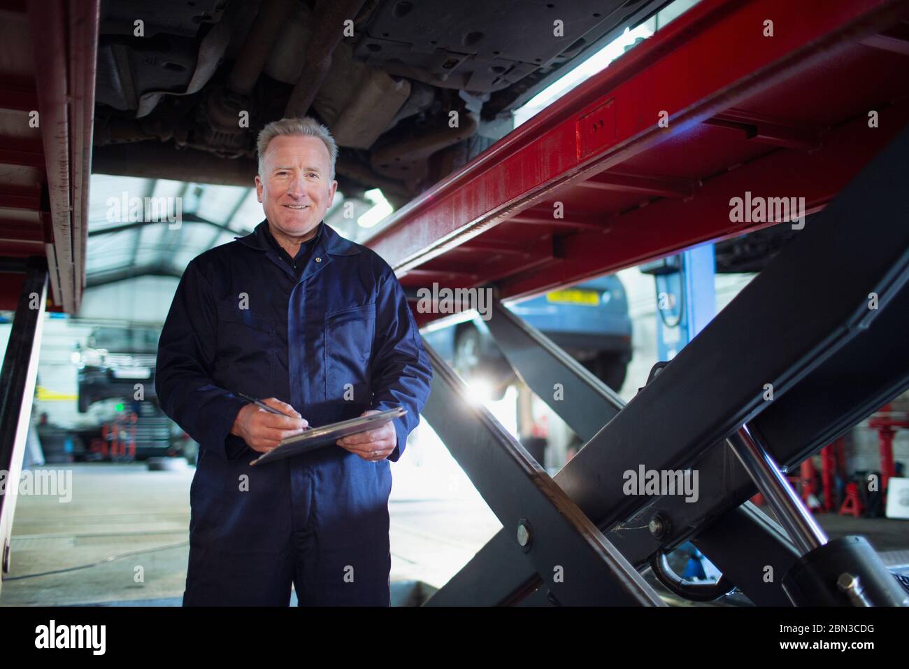 Portrait confident, smiling male mechanic with clipboard working under car in auto repair shop Stock Photo