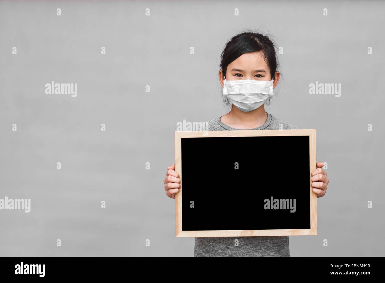 Little asian girl in a protective medical mask holding blackboard isolated on gray background. Protect from Coronavirus or Covid-19 epidemic. Stock Photo