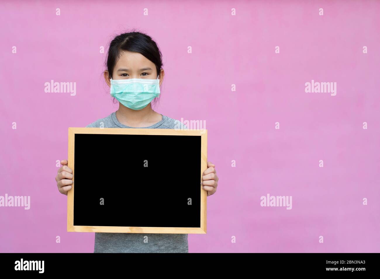 Little asian girl in a protective medical mask holding blackboard isolated on pink background. Protect from Coronavirus or Covid-19 epidemic. Stock Photo
