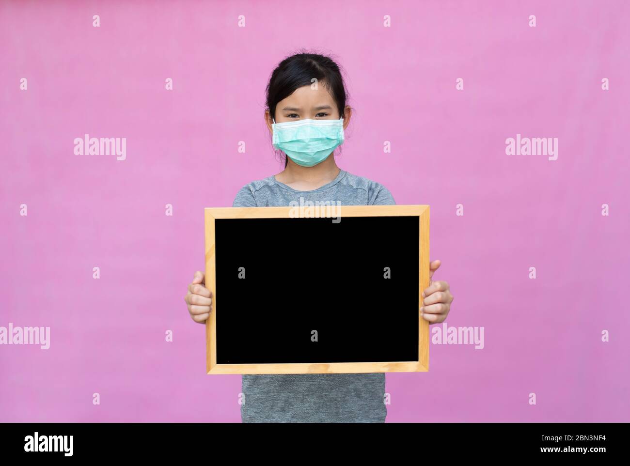Little asian girl in a protective medical mask holding black board isolated on pink background. Protect from Coronavirus or Covid-19 epidemic. Stock Photo