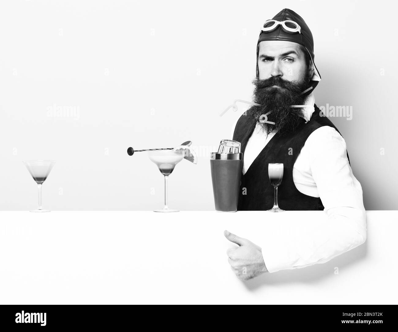 handsome bearded aviator or pilot with beard and mustache on serious face with shaker alcoholic cocktails in vintage suede leather waistcoat with hand and glasses, on purple green studio background Stock Photo