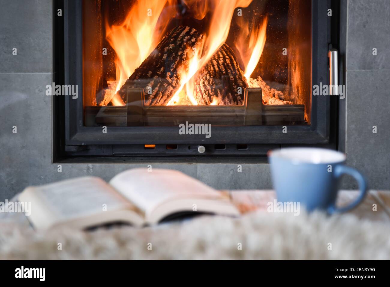 Open book, cup of tea and warm plaid near burning fireplace. Hygge concept Stock Photo