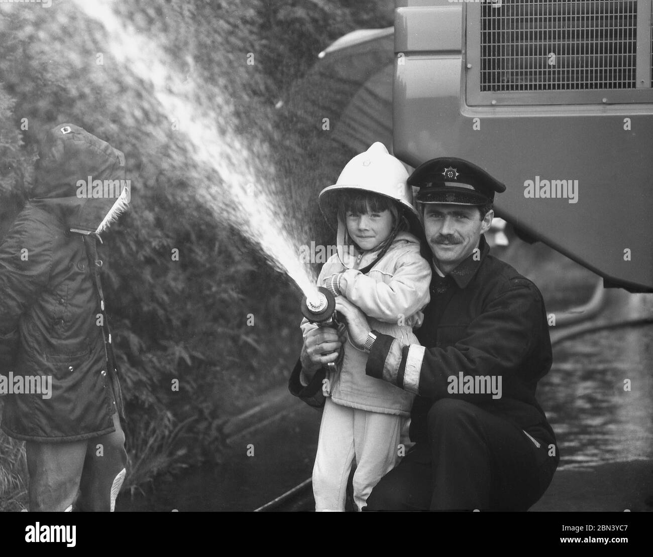 1980s, held by a uniformed fireman and watched by her brother, a little girl wearing a pointed fire helmet of the era, using a powerful water hose, at a fire service open day, England, UK. Stock Photo