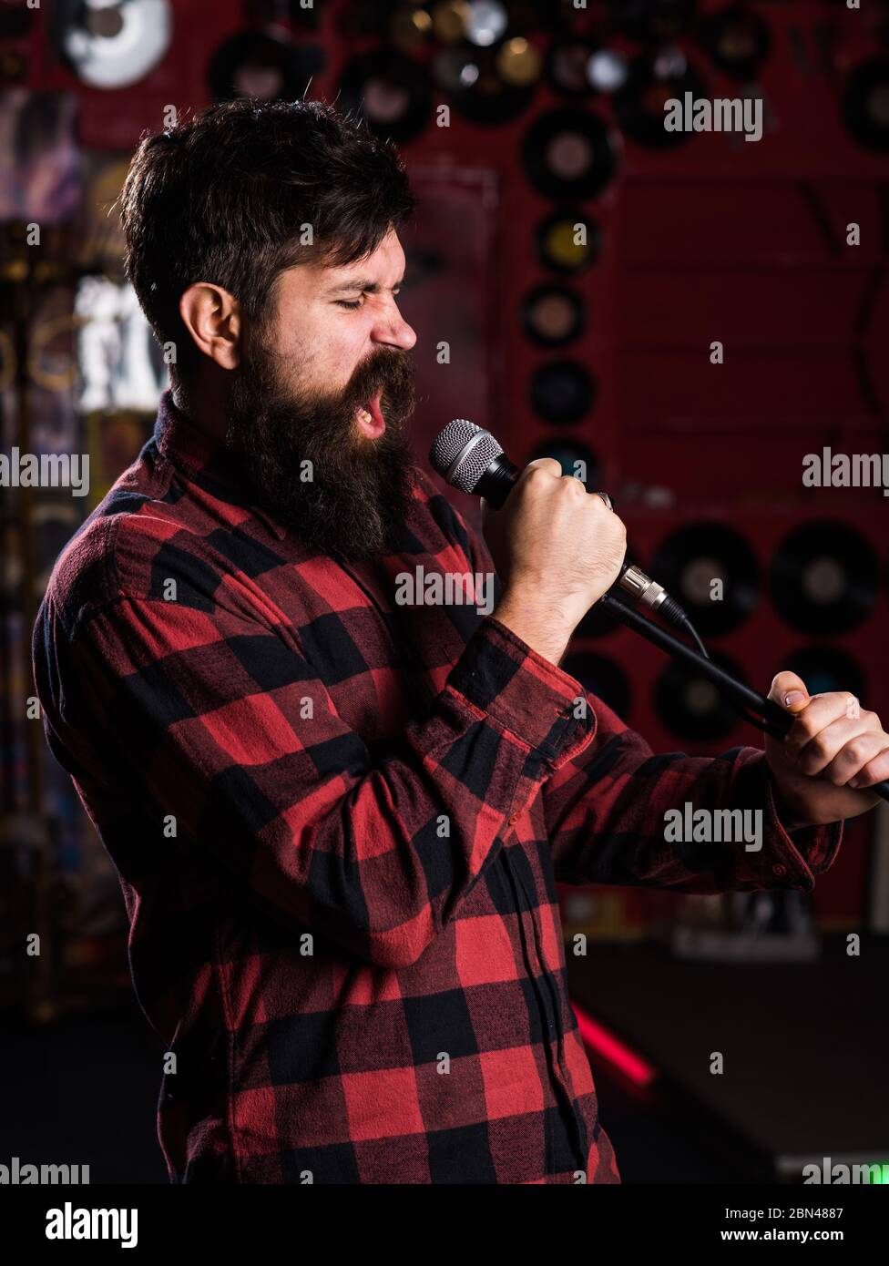 Man with enthusiastic face holds microphone, singing song, karaoke club  background. Musician with beard and mustache singing song in karaoke. Music  and leisure concept. Hipster likes to sing on stage Stock Photo -