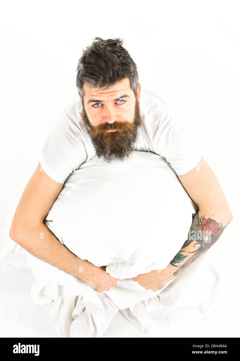 bearded guy waking up in morning sitting on bed in white shirt. Tattooed hipster looking up at camera. Top view. Stock Photo