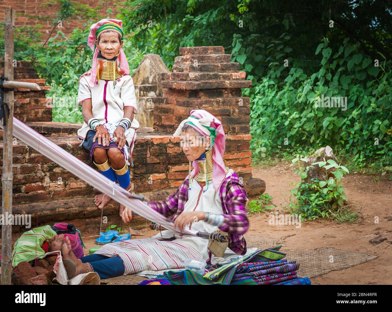 Bagan Myanmar - October 29 2013; Two Kayan women with brass rings around necks one weaving while other in selective focus sitting wall looks on. Stock Photo