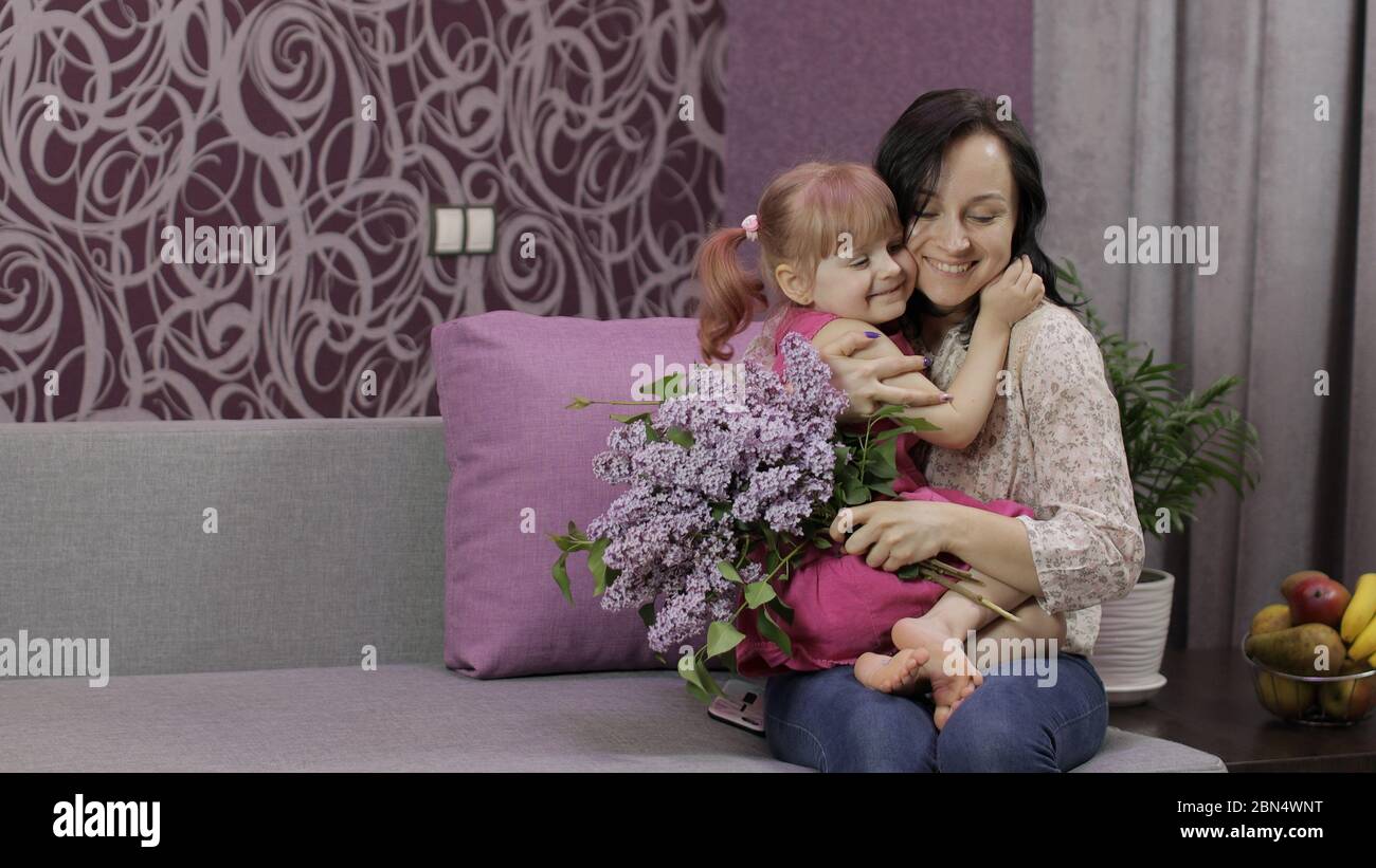 Little daughter and mother sitting on sofe with bouquet of lilac purple flowers at home. Little girl child hugs her mom, smiles. Kissing and hugging. Happy childhood, family. Mom and baby kid Stock Photo