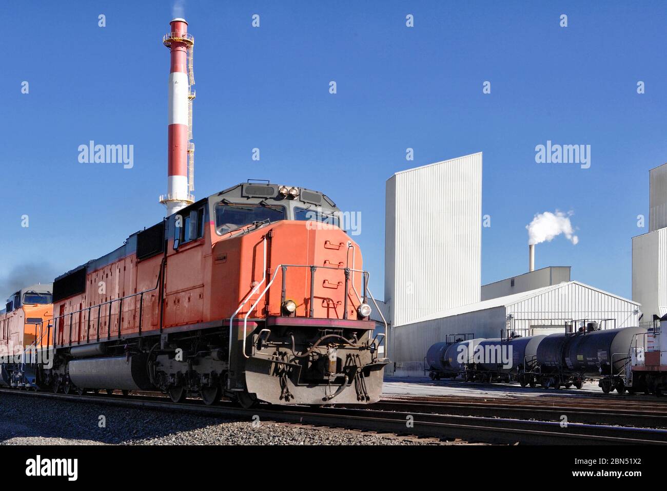 Train locomotives passing the smoke stack of an industrial manufacturing facility on a sunny day. Stock Photo