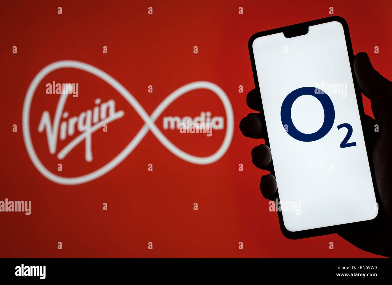Stone / United Kingdom - May 12 2020: O2 Telefonica logo on smartphone screen hold in hand and Virgin Media logo on blurred background. Concept for Vi Stock Photo