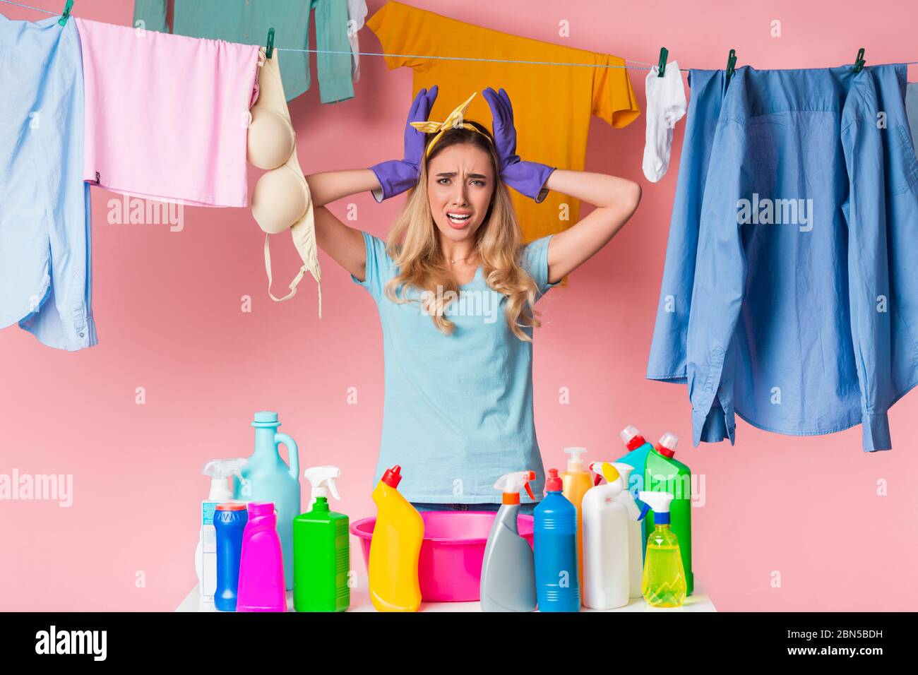 Sad housewife holds head with hands, in front of her is basin and cleaning supplies, behind is clean clothes on rope Stock Photo