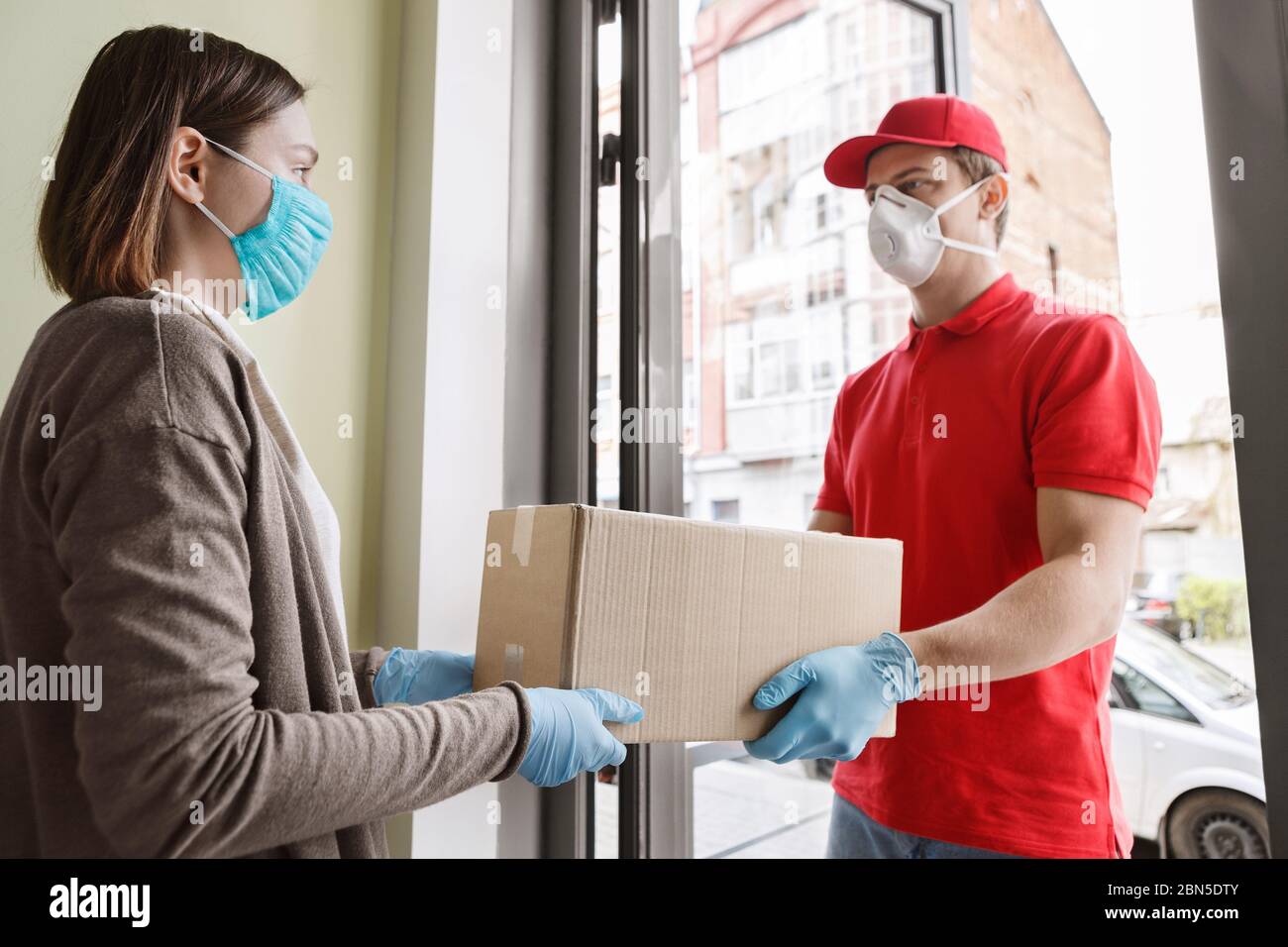 Courier delivery during quarantine. Girl opens door and takes parcel from deliveryman Stock Photo