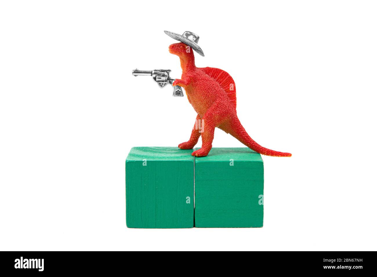 Toy tyrannosaurus rex wearing a cowboy hat and holding a revolver in his arm stands on two green wooden blocks. Close-up shot, isolated on white. Stock Photo