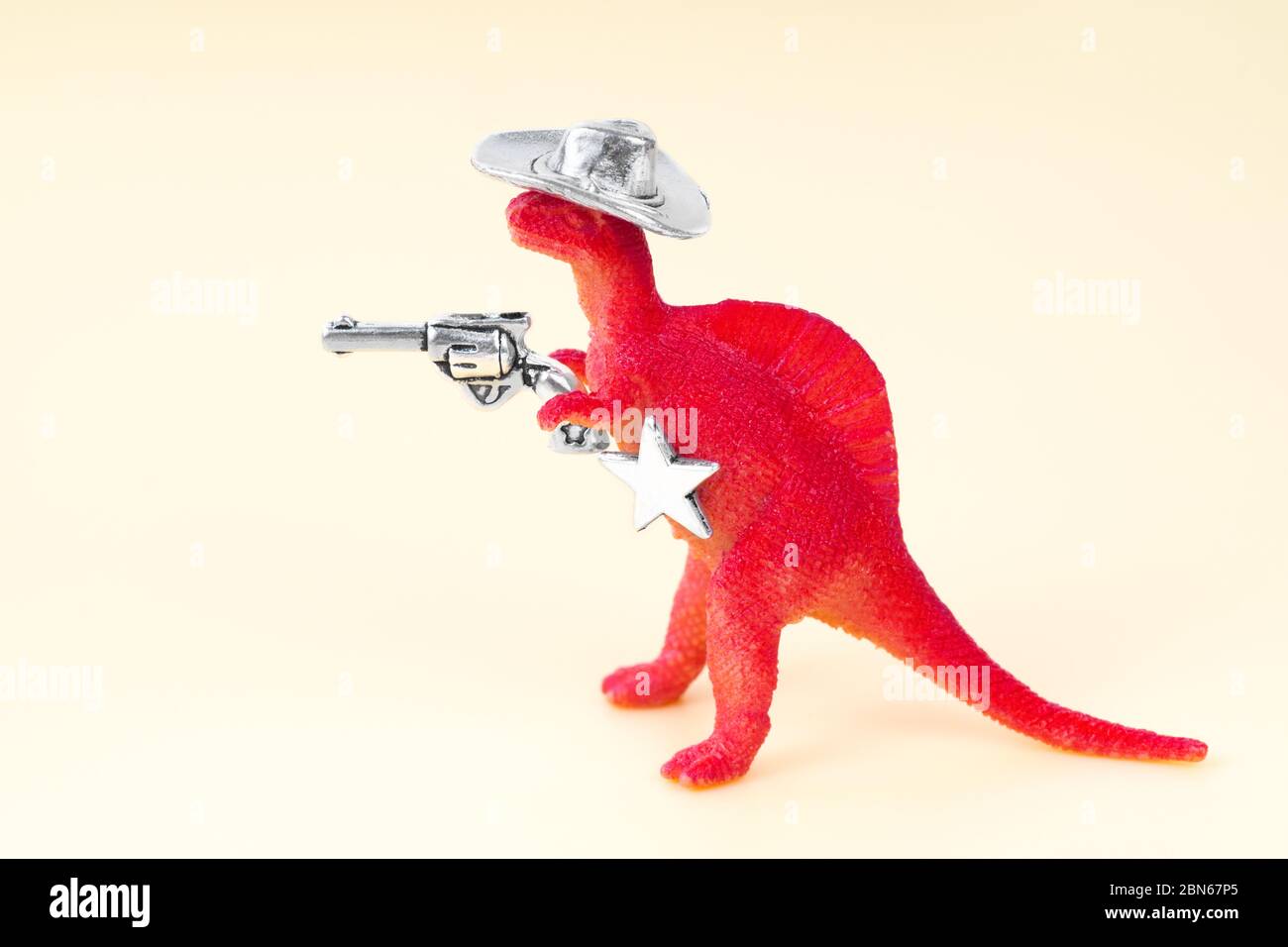 Close up of a small red toy dinosaur wearing a cowboy hat, holding a revolver in his arm and having a sheriff star on a neutral background. Stock Photo