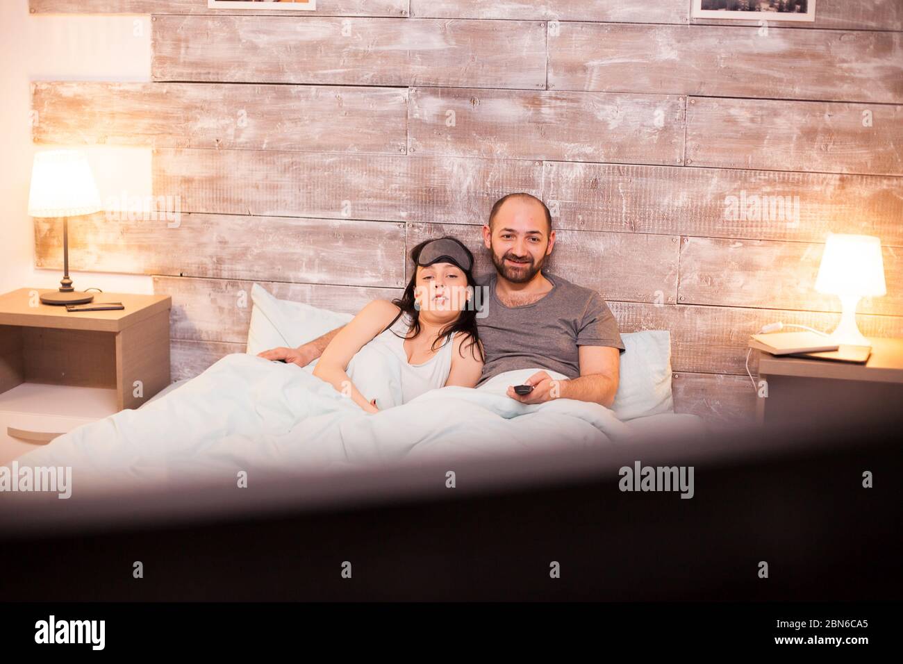 Young couple in pajamas watching tv lying under the blanket on