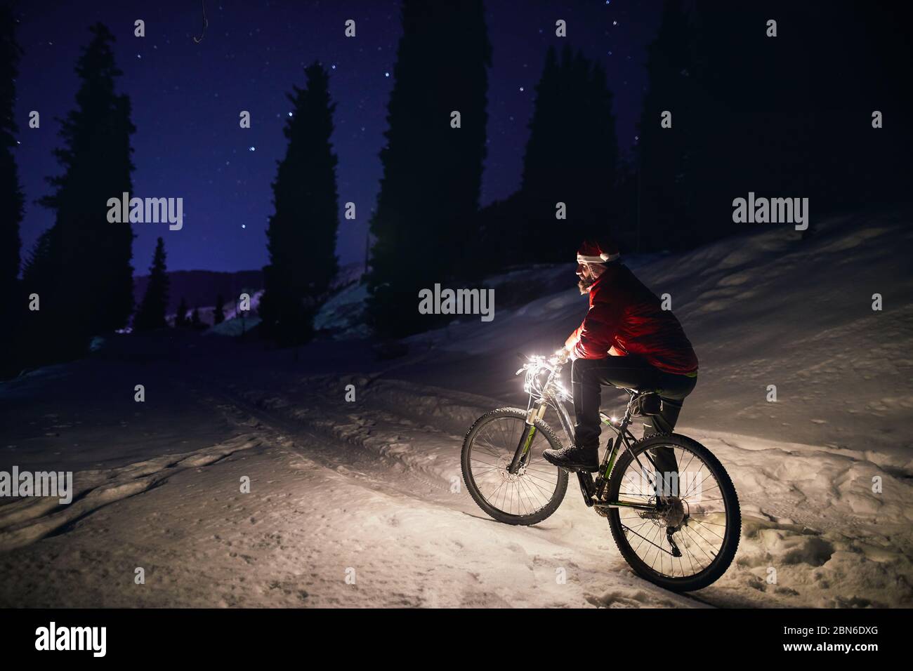 Man in red jacket with bicycle and Christmas hat at winter snowy forest in the mountains under night sky with stars Stock Photo