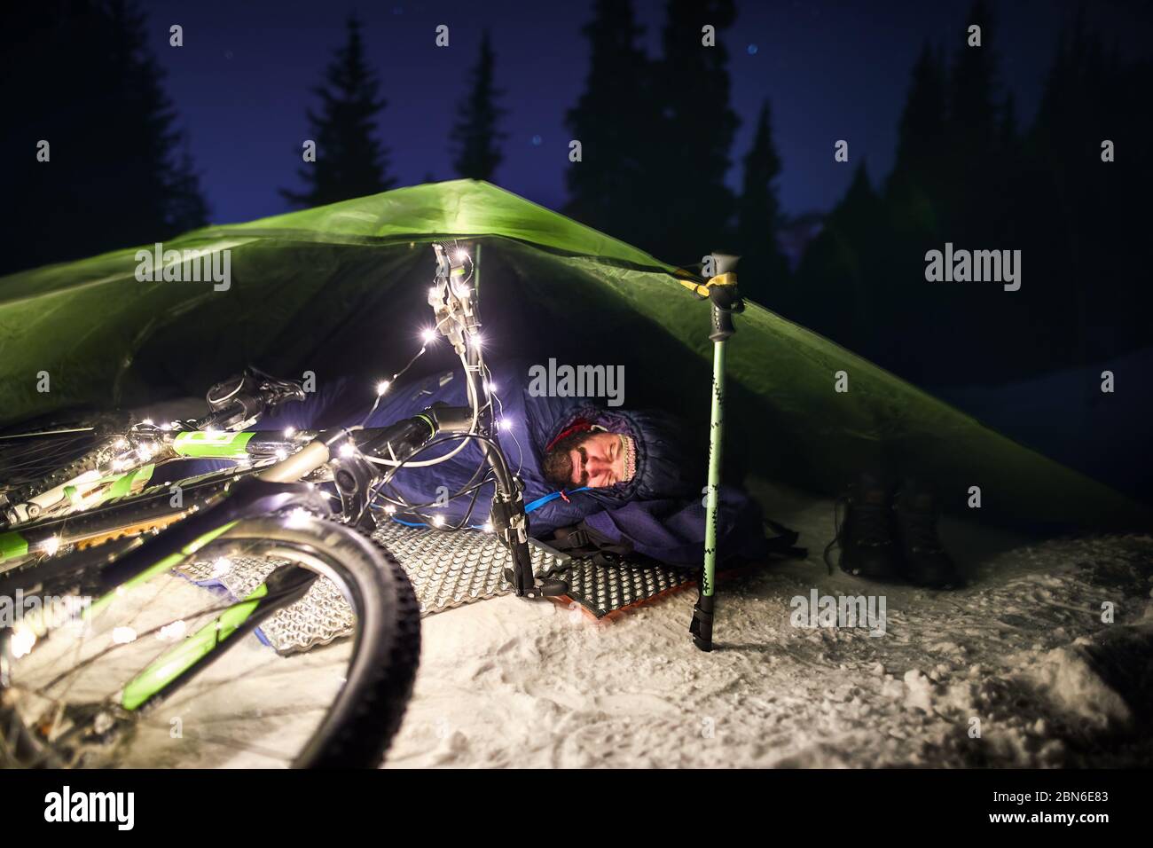 Bearded man is sleeping in tent at sleeping bag with bicycle at winter night. Handlebar decorated with Christmas lights. Bikepacking and Christmas con Stock Photo