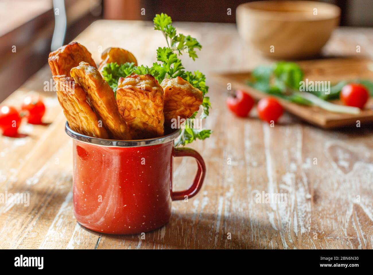 Red enameled cup with fried tempeh (traditional Indonesian soy product) on wooden surface. Side view. With space. Stock Photo