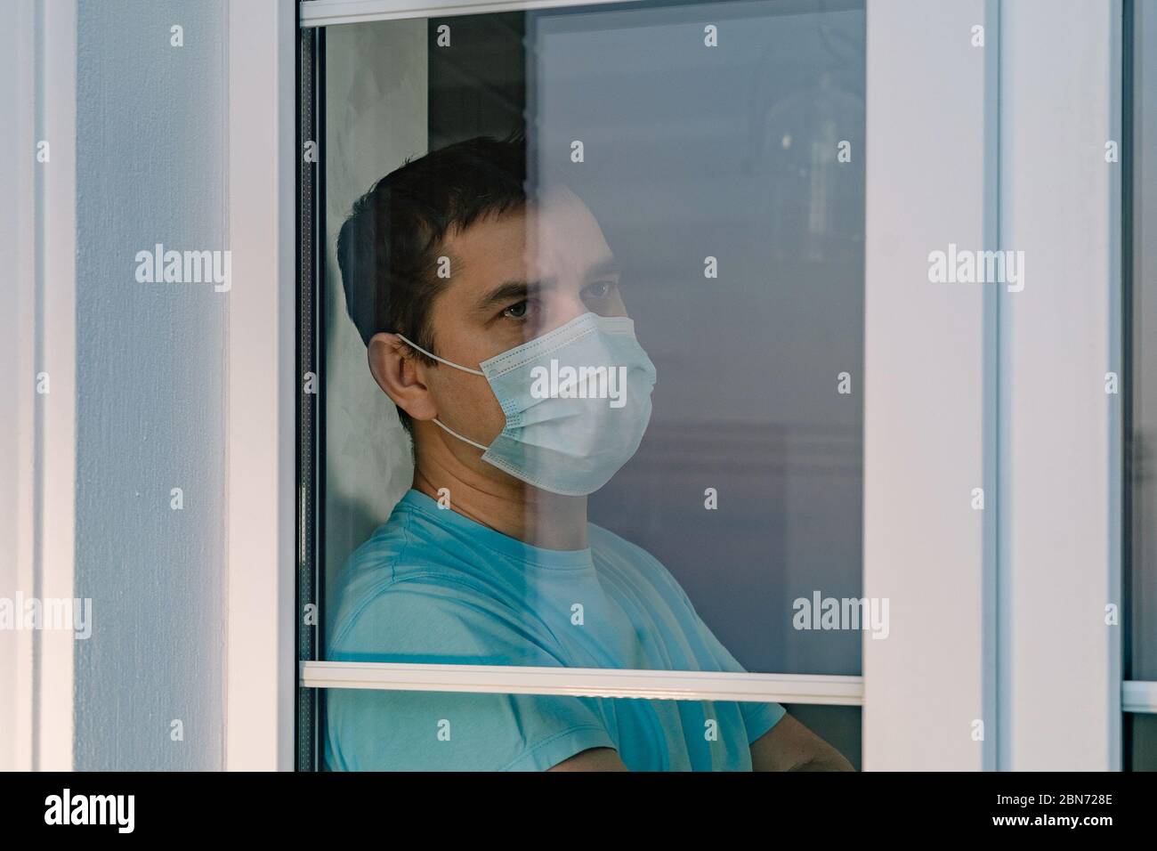 Man in medical mask looks out window, COVID-19 Stock Photo