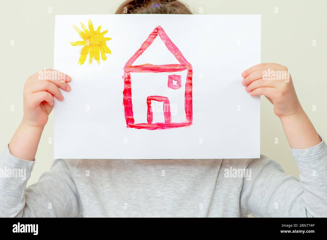 Child's hands holding picture of house with sun covering her face on yellow background. Childs with drawn house. Stock Photo