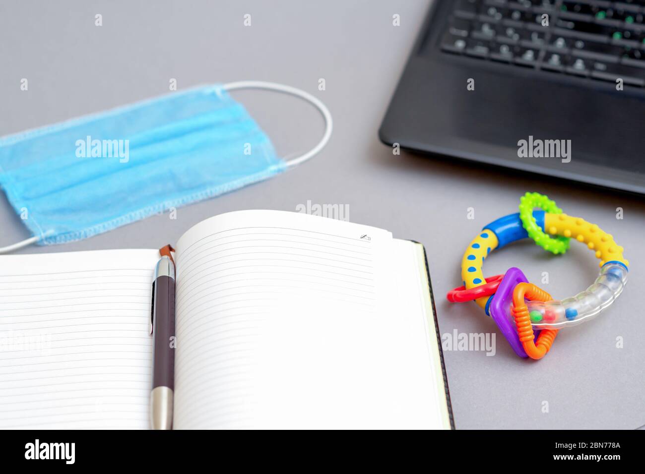 Protective medical mask near the laptop and pen with open notebook and rattle toy on gray background. Concept of working from home. Stock Photo