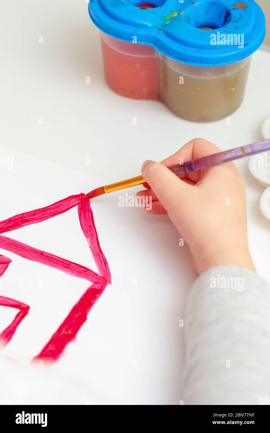 Closeup of child's hand with brush drawing the house on white paper by watercolors. Stock Photo