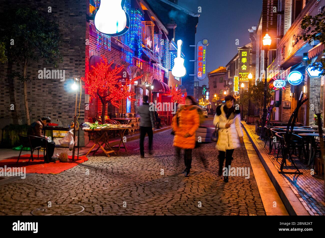 Three ladies walk along a cobbled pedestrian street. A man waits for customers at his barbecue stand in Laowaitan, Ningbo Stock Photo