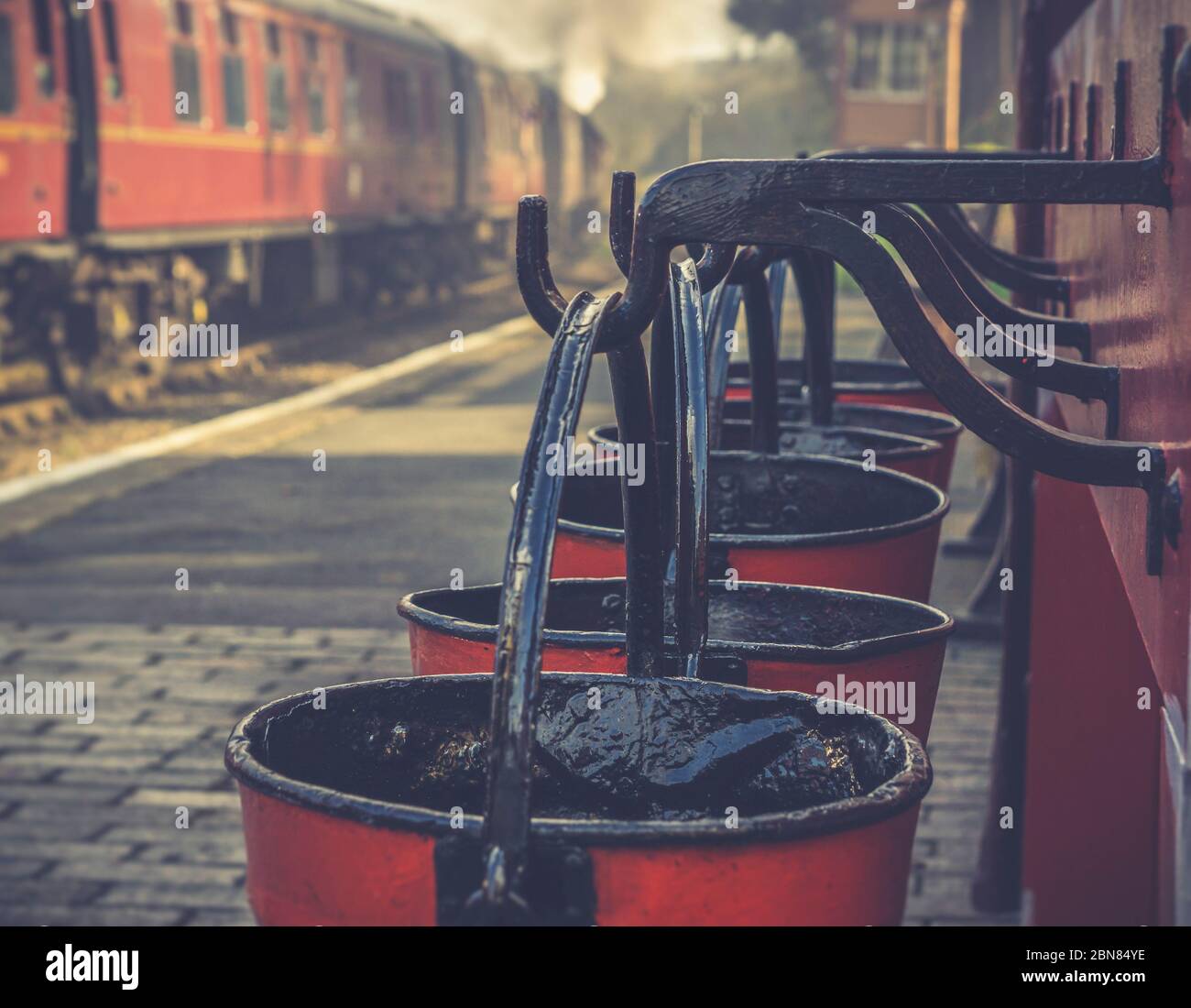 Retro close up of hanging vintage fire buckets on platform of heritage railway station, Severn Valley Railway UK, vintage steam train is leaving. Stock Photo