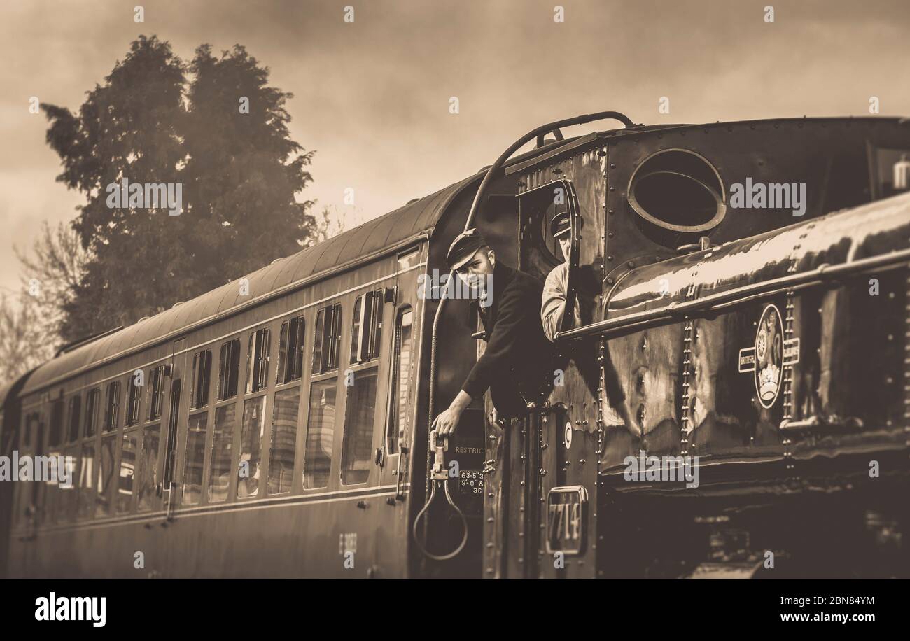 Moody, sepia side view of vintage UK steam train approaching, steam locomotive fireman cab crew prepare to catch railway token from station signalman. Stock Photo