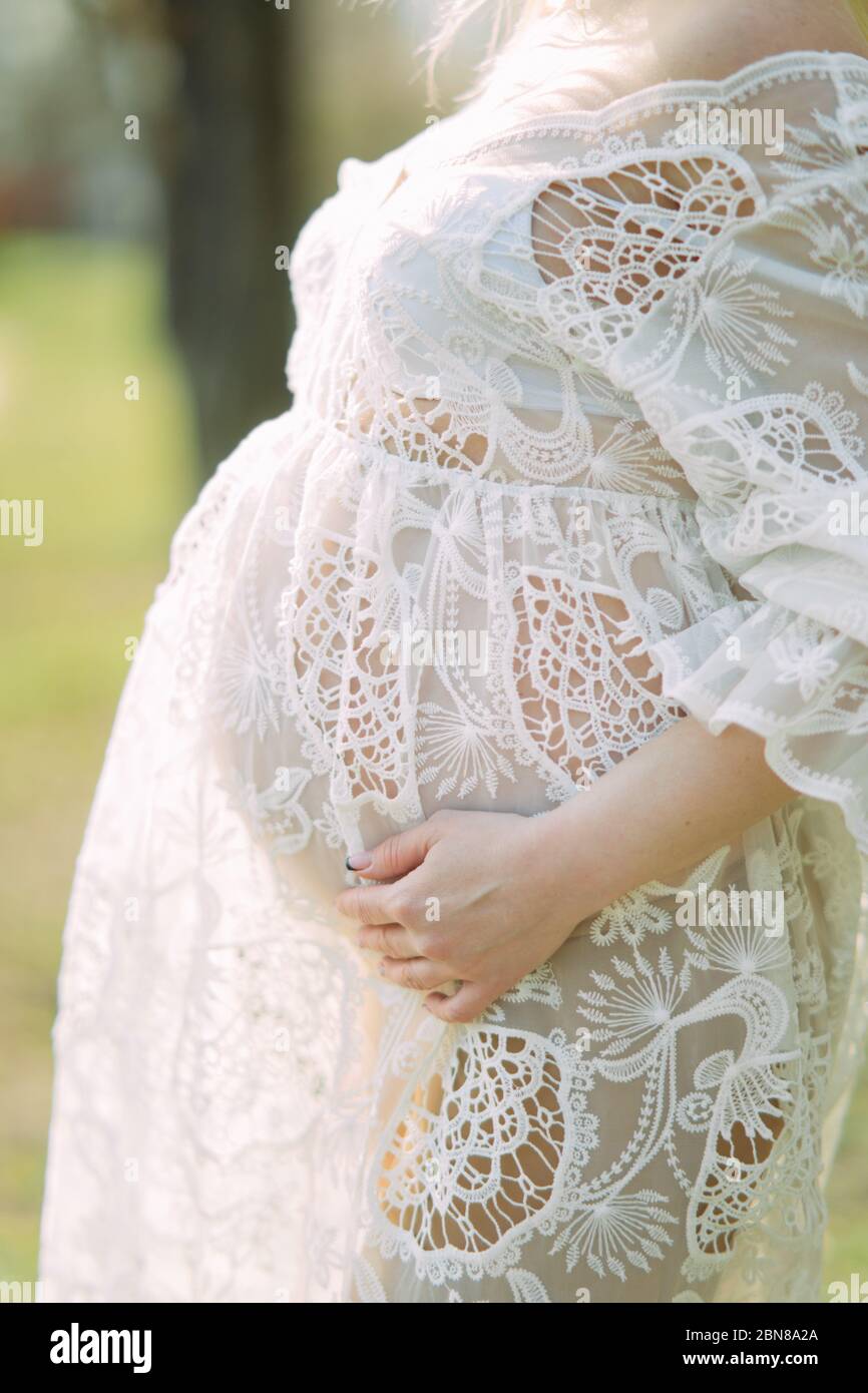 Pregnant woman stands in white lace dress and supports her belly with ...