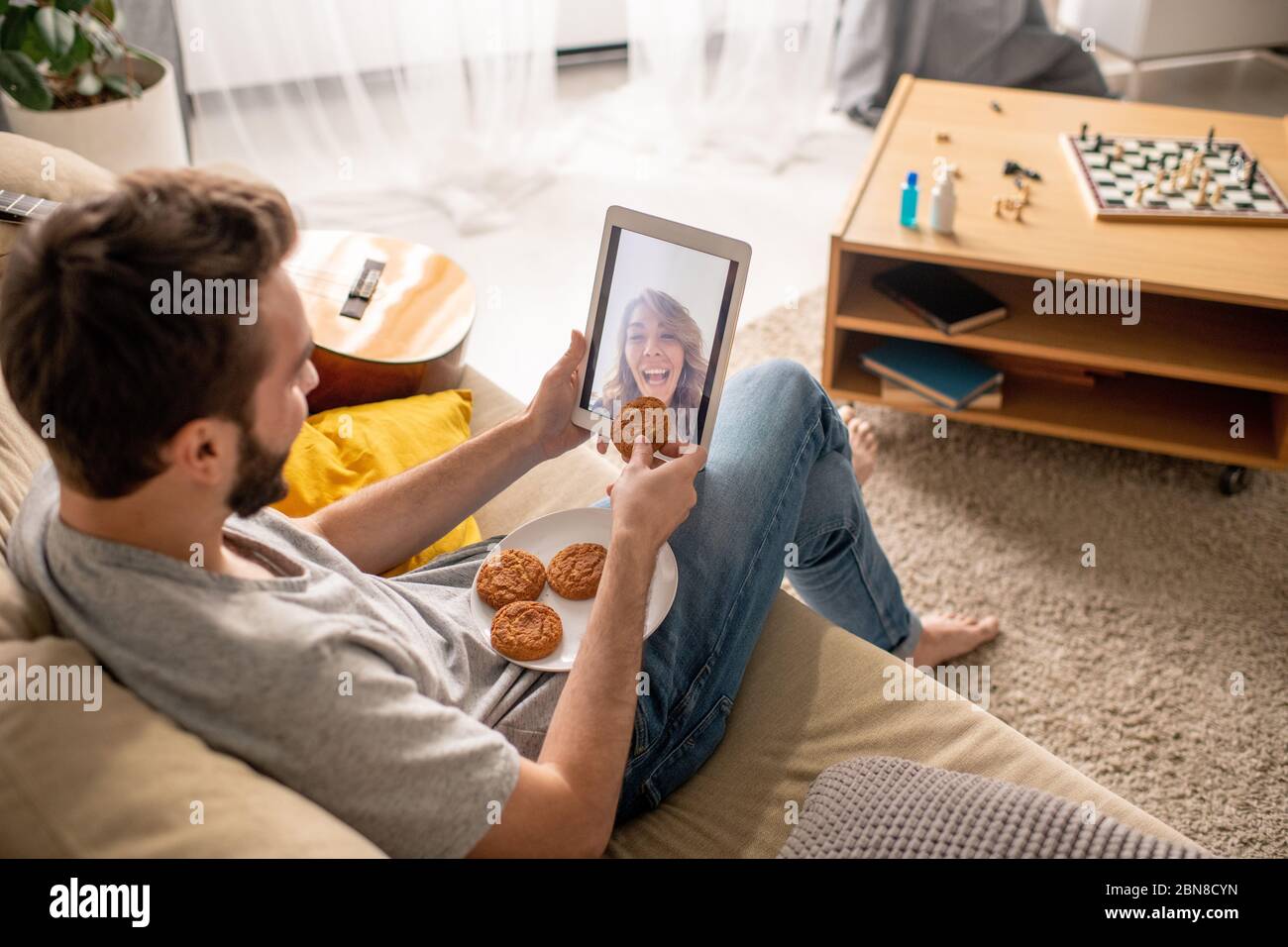 Young man sitting on sofa and using tablet while chatting with girlfriend, she pretending to eat cookie given by him through tablet screen Stock Photo