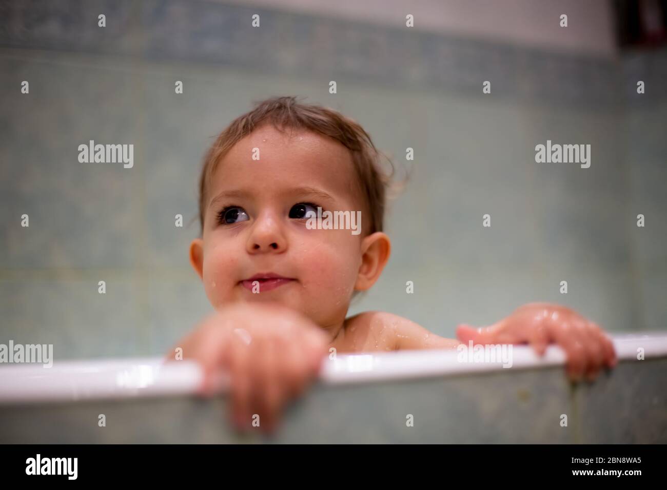 A cute Caucasian baby peeks out of the bathtub, put hands resting on