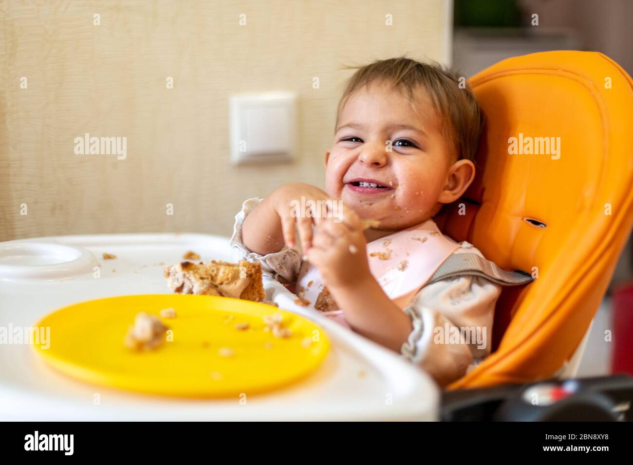cute baby sits in an orange baby chair with a table with dirty hands and face, looks in camera and laughs. on table are crumbs and an orange plate Stock Photo