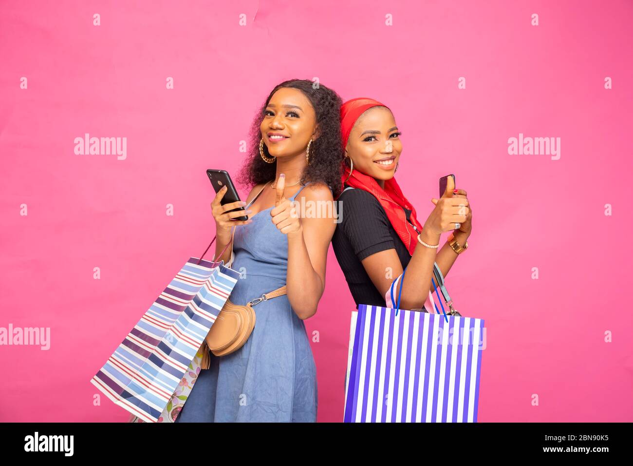 beautiful young african girls holding shopping bags standing back to back, holding their phones, smiling and giving thumbs up Stock Photo