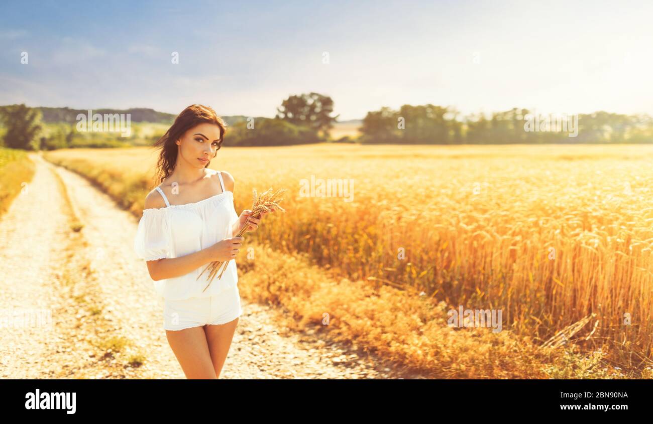 Beauty romantic girl enjoying nature in outdoors. Happy young woman in white shorts holding the ears on the road near of golden ripe wheat field in su Stock Photo